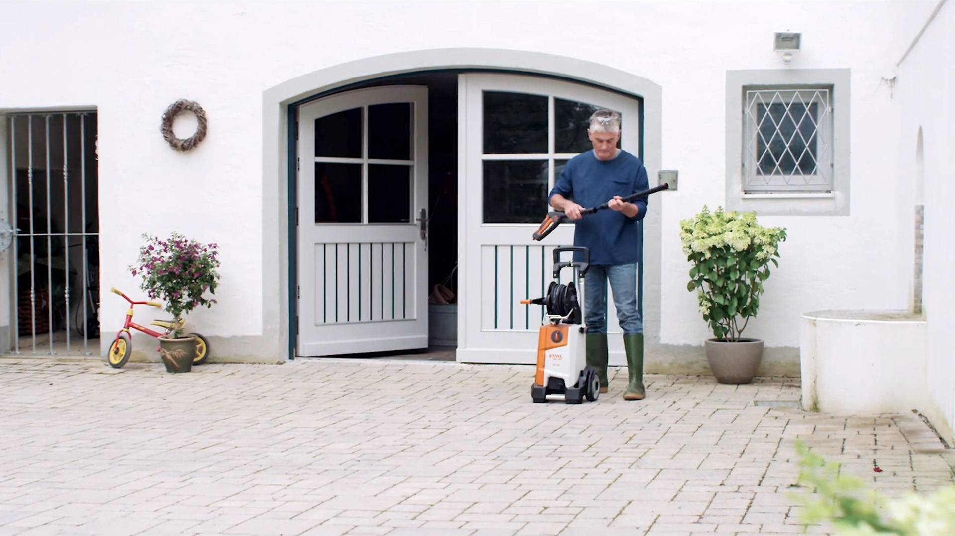 Um homem à frente de uma casa branca a preparar-se para limpar o exterior com uma máquina de limpeza de alta pressão STIHL