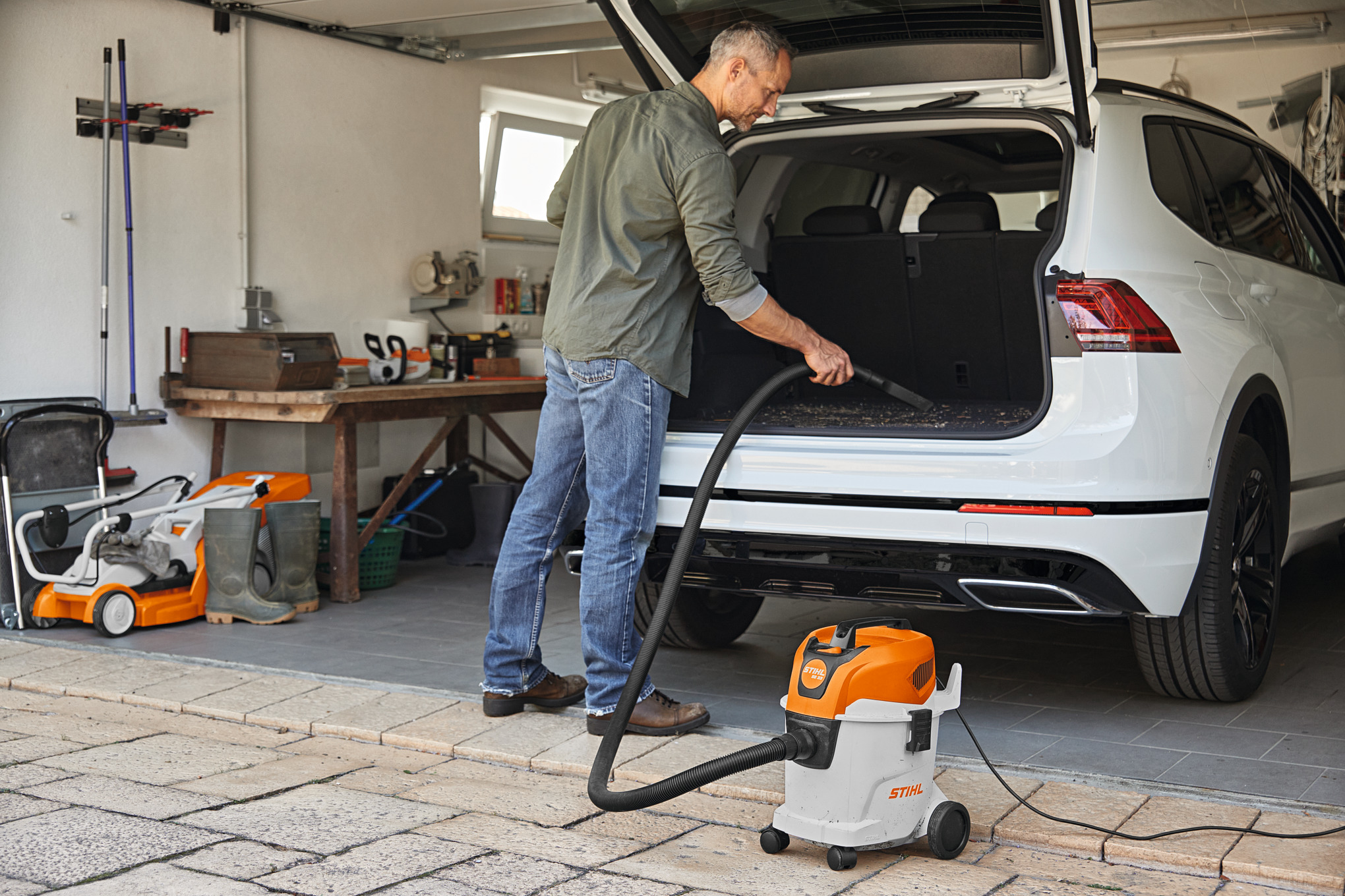 Homem limpa o interior do carro com um aspirador STIHL SE 33 na garagem de sua casa, onde também guarda outras máquinas, como motosserras e cortadores de relva.