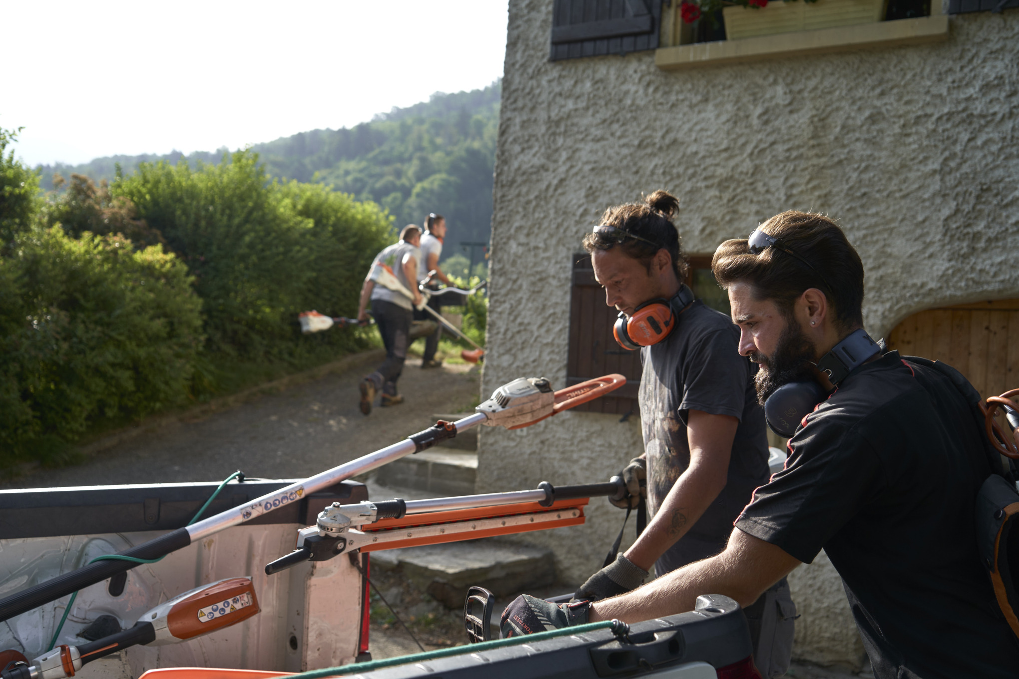 Dois jardineiros paisagistas descarregam máquinas a bateria STIHL de um carro, em segundo plano estão outros trabalhadores 