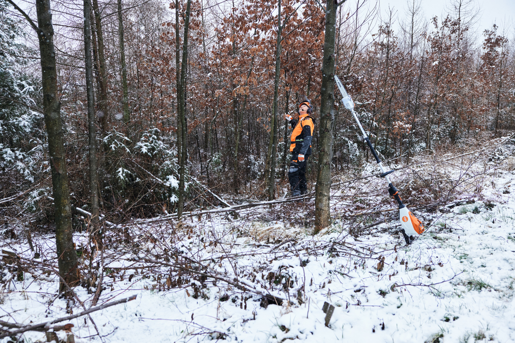 Um homem a trabalhar com equipamento de proteção individual numa floresta coberta de neve, em primeiro plano, uma podadora em altura a bateria STIHL HTA 150 encostada a um tronco de árvore