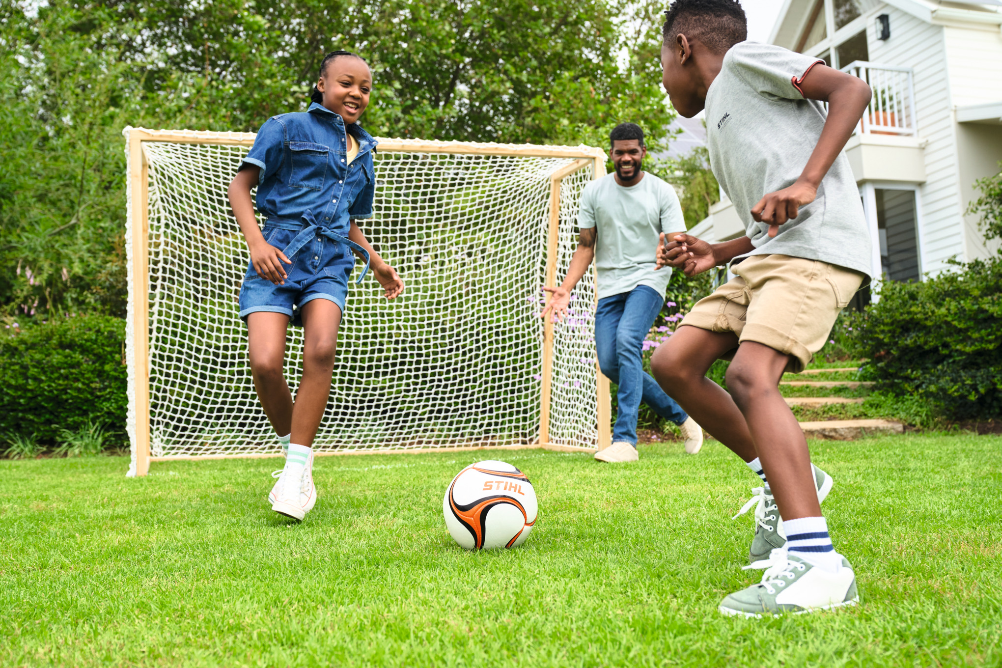 Três crianças jogam futebol num espaço verde com uma baliza de futebol em madeira