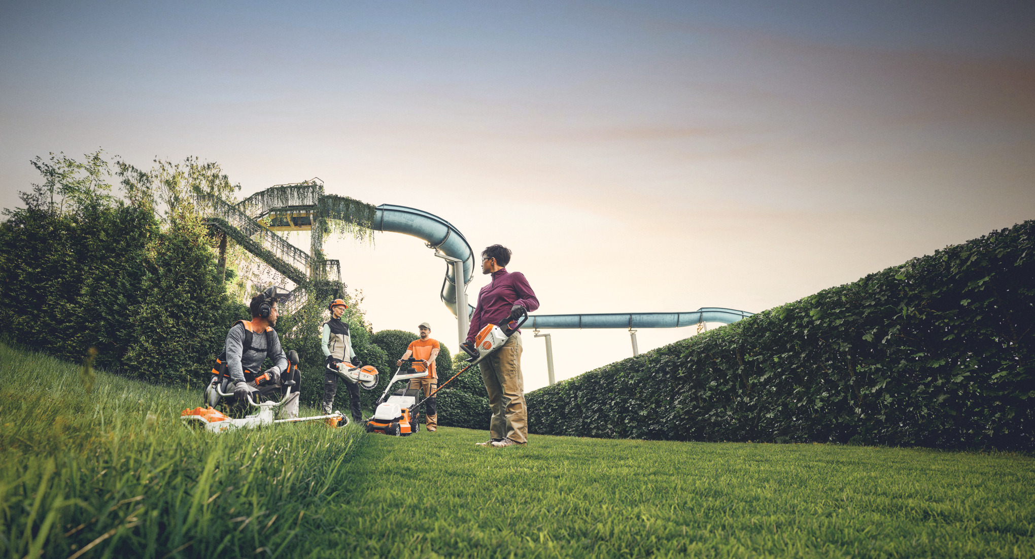 Um grupo de pessoas trabalha com máquinas de jardinagem STIHL num espaço verde, com uma grande sebe e escorregas de água em segundo plano. Estão equipados com foices a motor, corta-relvas e equipamento de proteção.