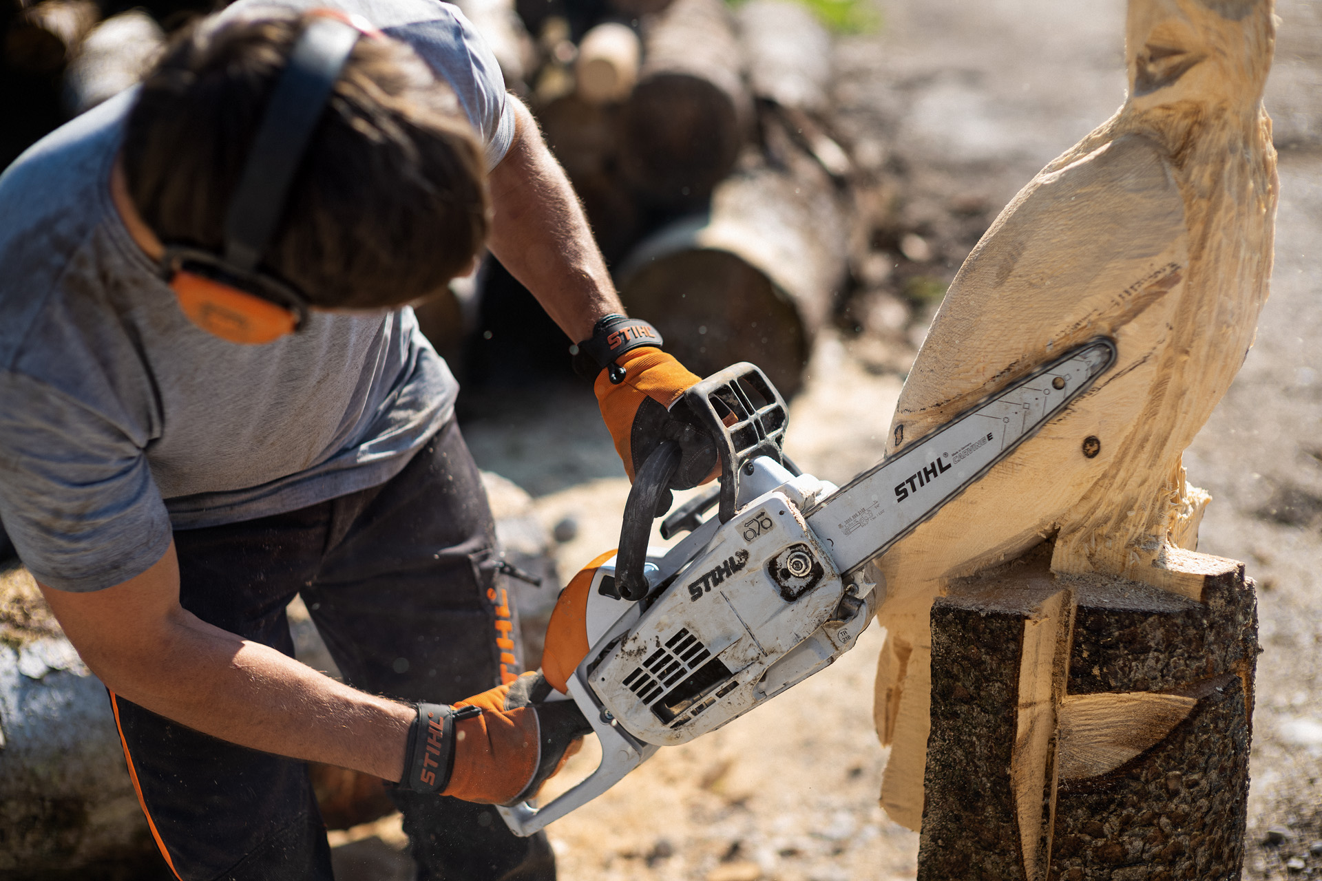 Homem com proteção auditiva e luvas usa motosserra de esculpir STIHL MS 193 C-E para trabalhar escultura de madeira.