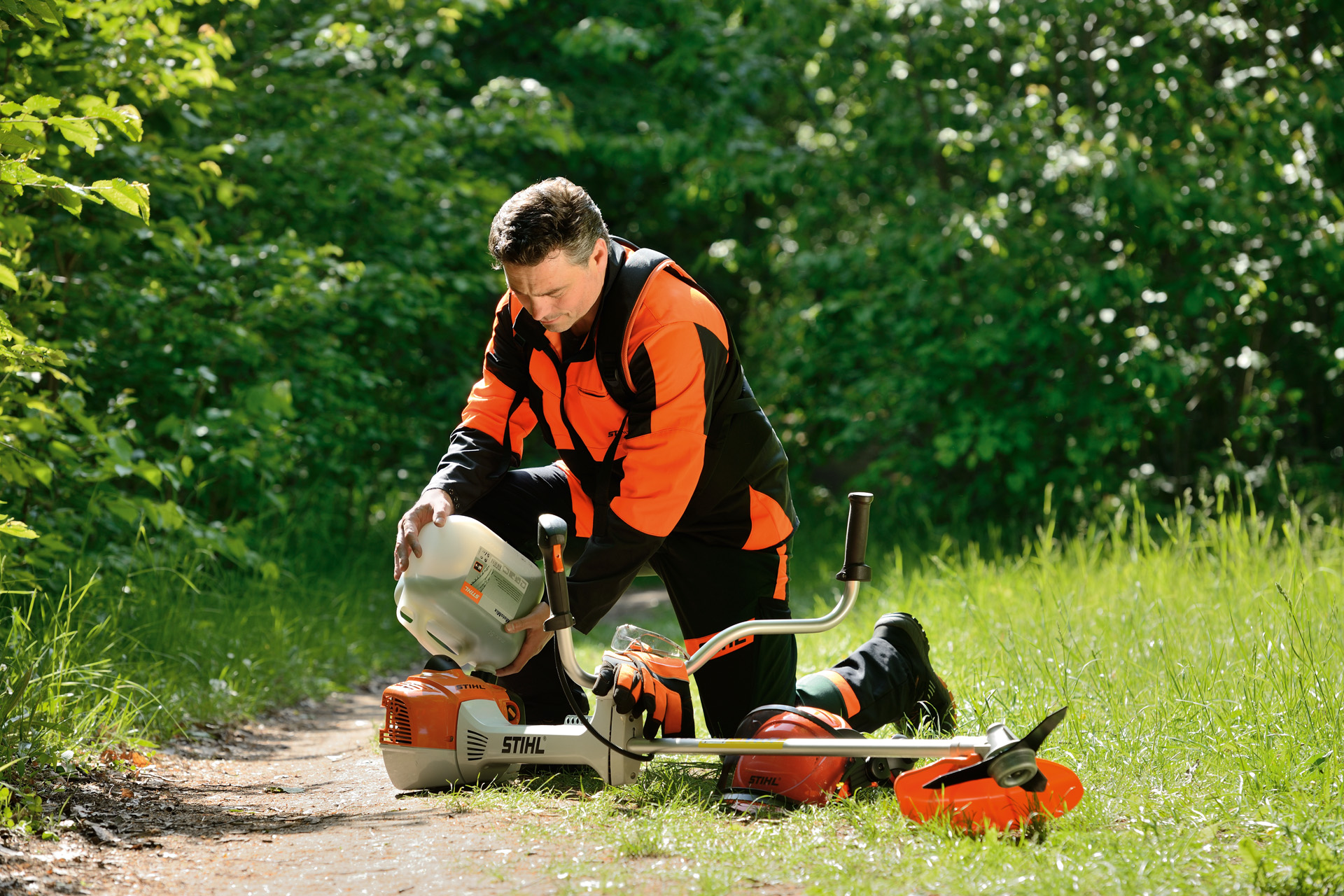 Um homem com um garrafão a abastecer o depósito de combustível de uma  motorroçadora STIHL com arbustos atrás dele