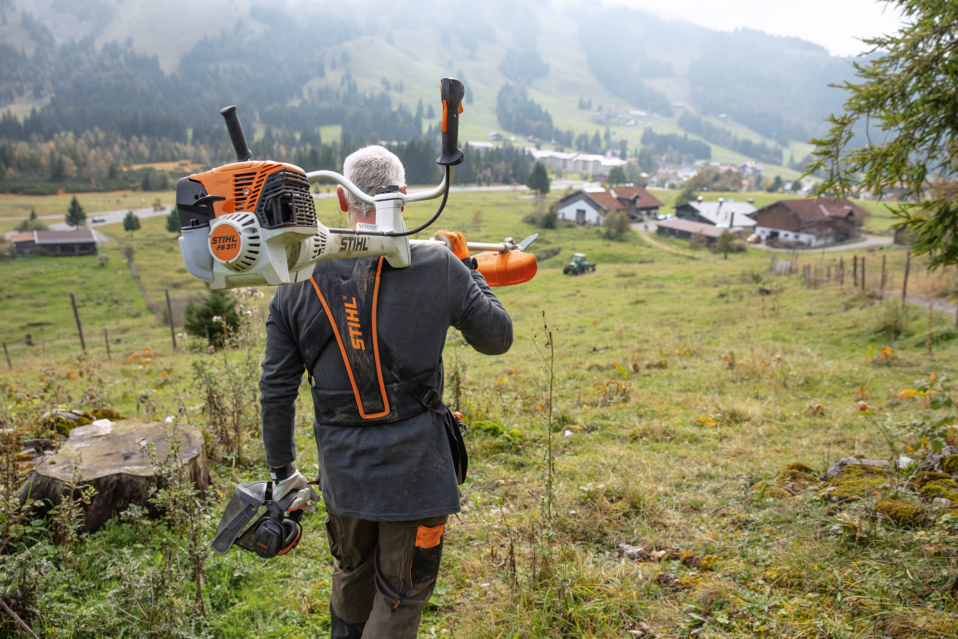 Um homem que segue as dicas para motorroçadoras com uma STIHL FS 311 ao ombro e uma colina verde e casas ao fundo