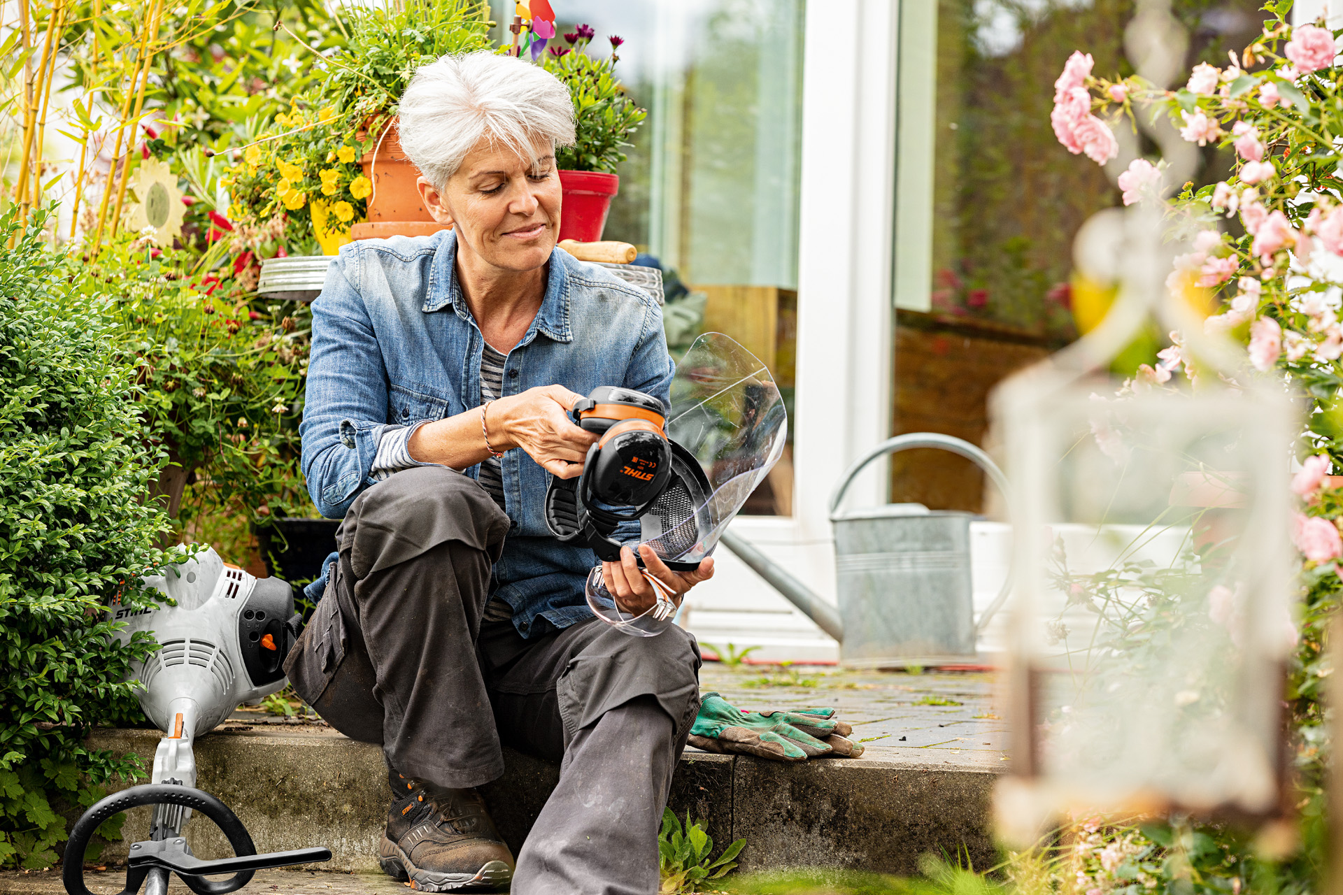 Uma mulher sentada no jardim, segurando uma viseira STIHL com protetores auditivos, ao pé de uma motorroçadora no chão.