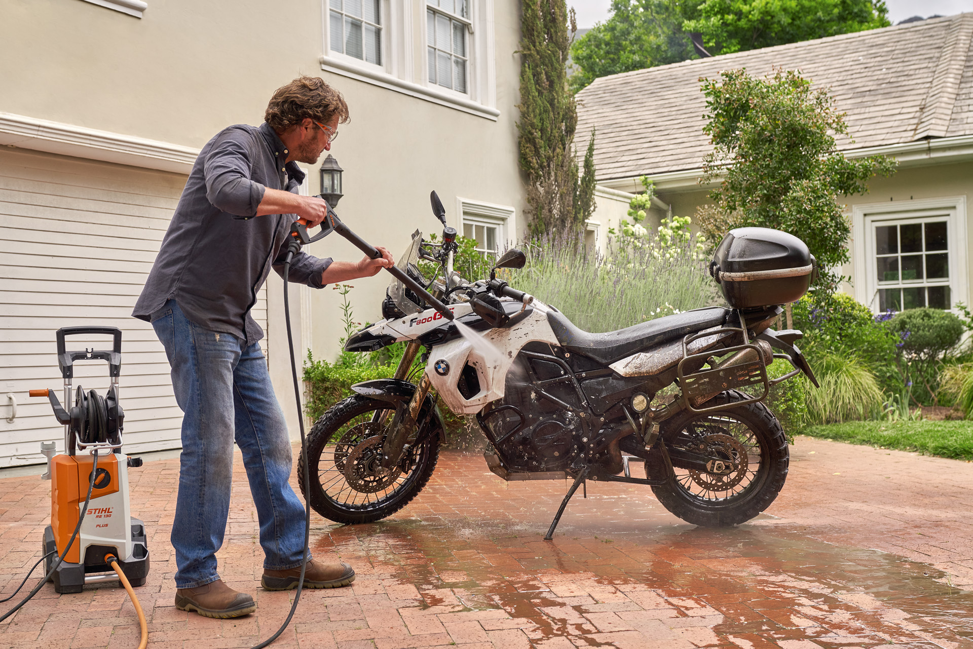 Homem a usar uma lavadora de alta pressão STIHL RE 130 Plus para lavar uma moto branca na entrada de uma casa