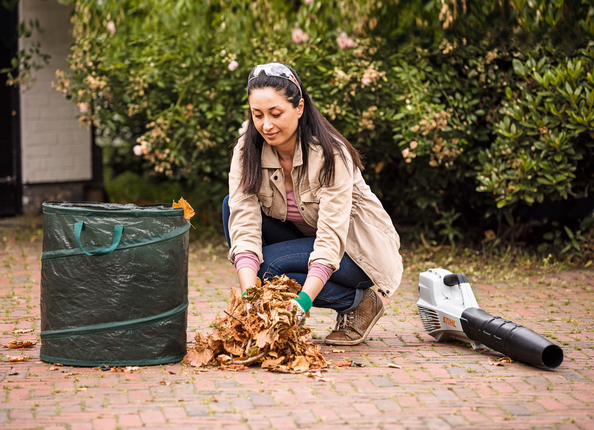 Uma mulher a limpar folhas e a encher um saco, tendo um soprador STIHL BGA 56 pousado no chão junto a ela