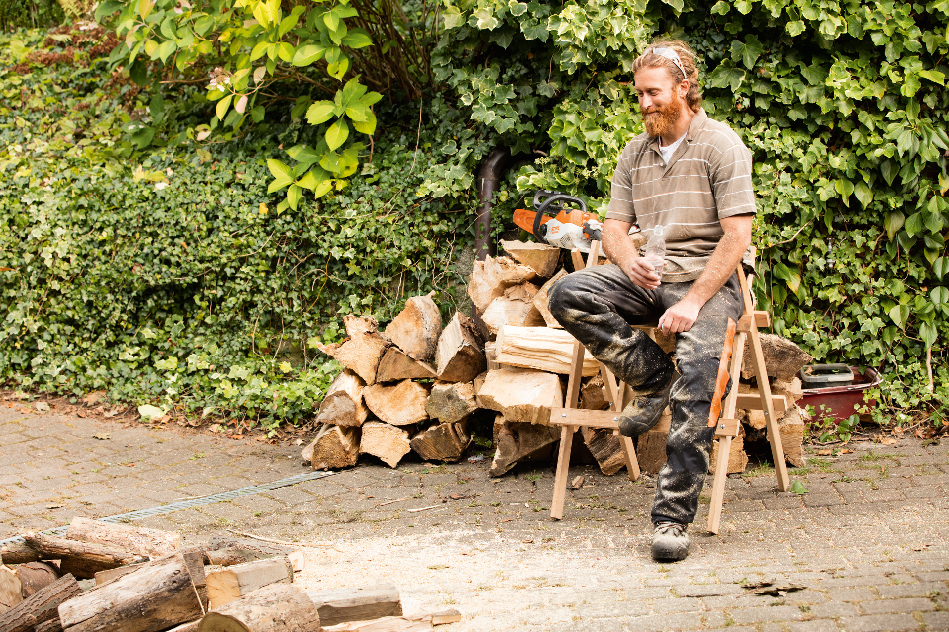 Homem sentado num cavalete à frente de uma pilha de troncos serrados.