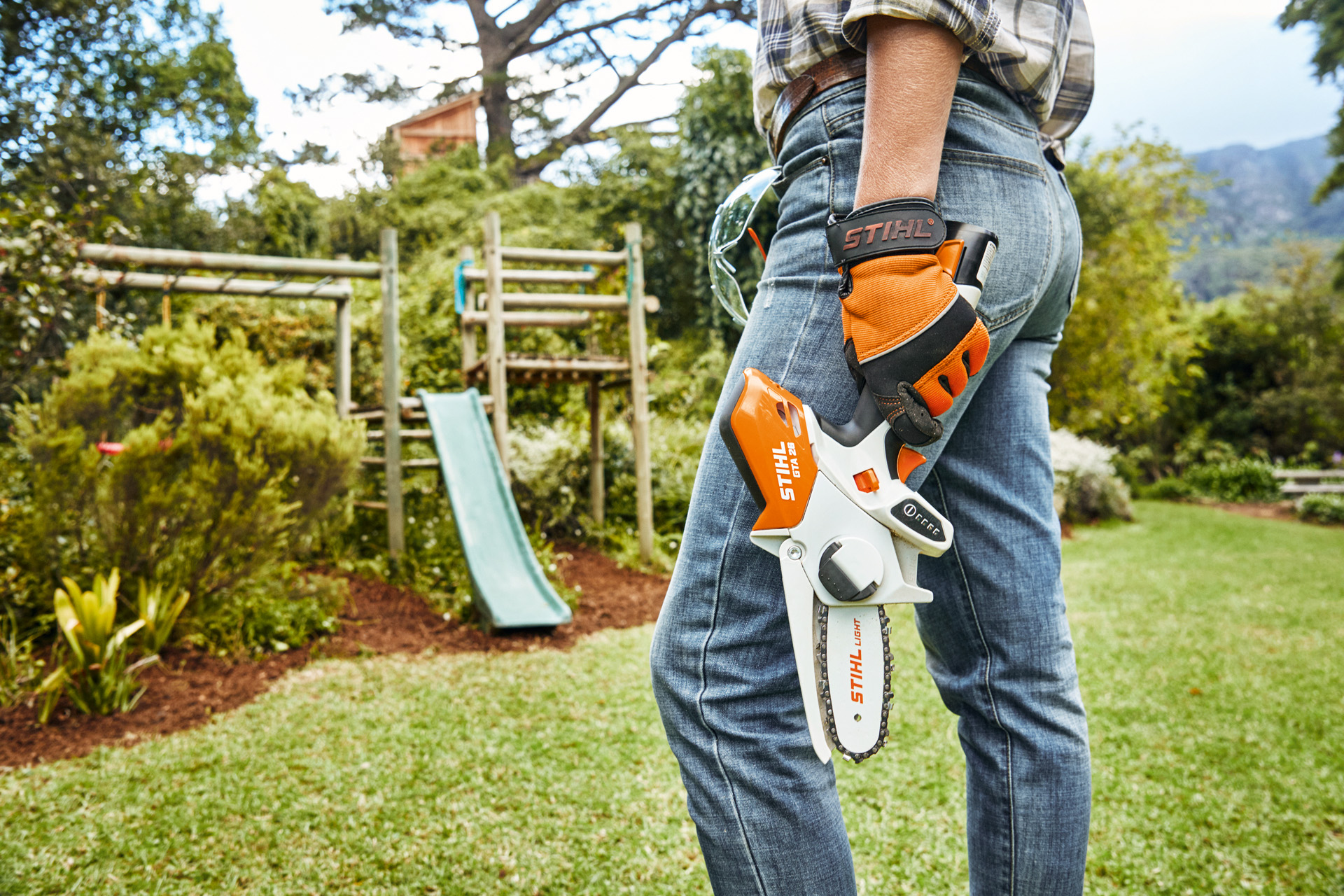 Uma pessoa com uma minipodadora de corrente a bateria STIHL GTA26 em frente a um escorrega no espaço verde
