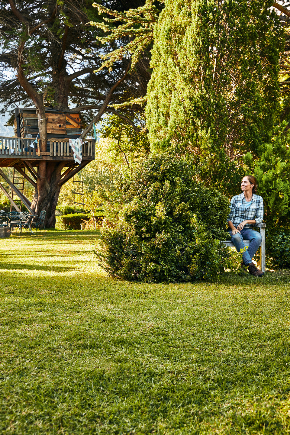 Uma mulher sentada num banco de jardim com arbustos e relvado e uma casa na árvore em segundo plano