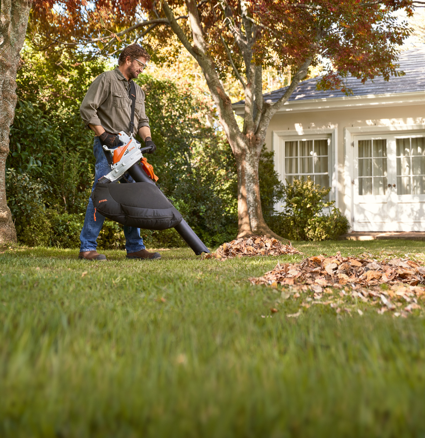 Homem a aspirar folhagem em frente a uma árvore no espaço verde, com um aspirador/picador a bateria STIHL SHA 56