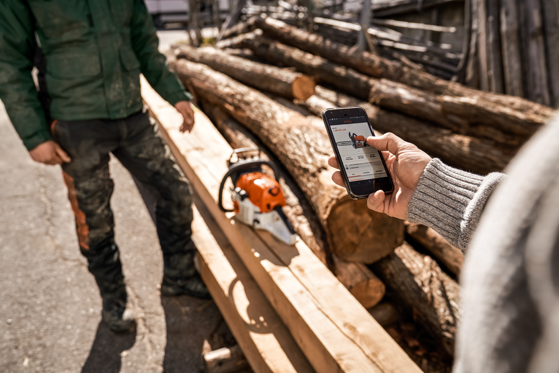 A aplicação STIHL connected em utilização num smartphone, com uma motosserra sobre uma pilha de madeira em segundo plano