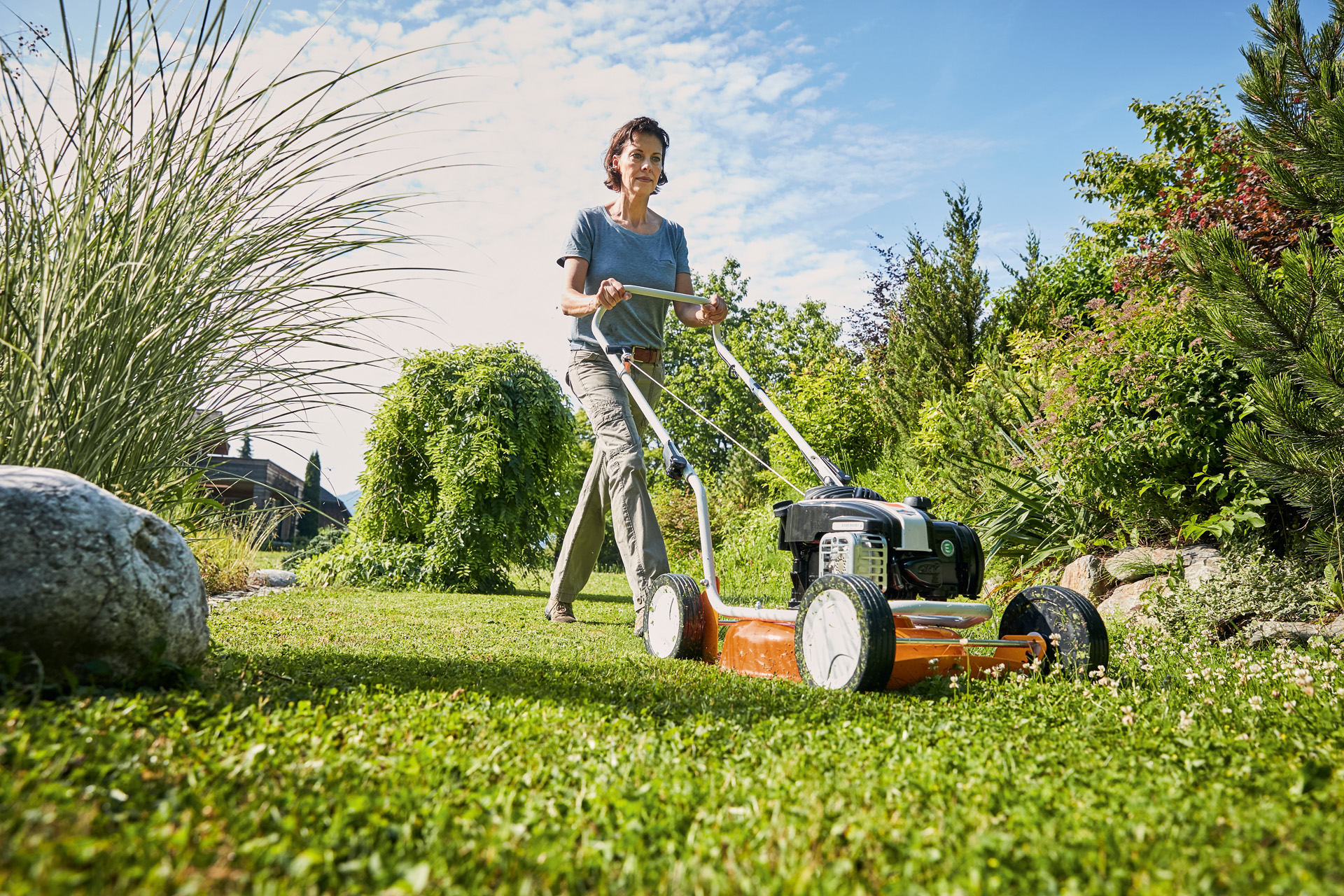 Uma mulher a utilizar um corta-relva com mulching a gasolina STIHL RM 2 R num relvado rodeado de arbustos e gramíneas