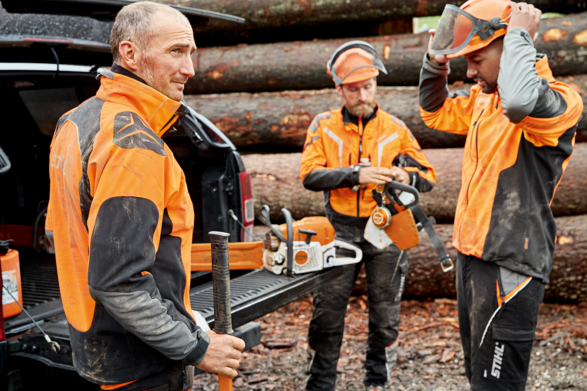 Segurança no trabalho: três homens com vestuário de proteção STIHL preparam-se para trabalhar, com uma motosserra STIHL