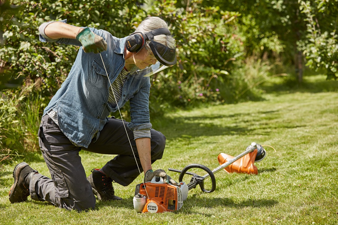 Uma mulher com proteção auditiva e facial liga uma motorroçadora a gasolina FS 56 R da STIHL num terreno verde