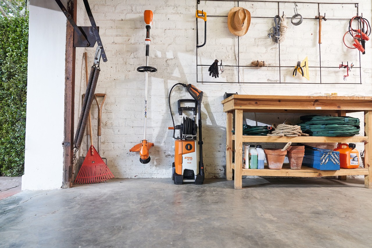 armazenamento de uma máquina de limpeza de alta pressão STIHL RE 130 PLUS e de uma motorroçadora STIHL numa garagem, junto a outros utensílios para o jardim